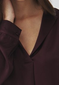 Burgunderfarbene Bluse mit tiefem V-Ausschnitt und glatter Textur. Verfügt über einen knöpfbaren Manschettenverschluss und leichte Drapierungen am Körper.