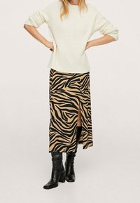 Cremfarbener Strickpullover mit umgeschlagenen Ärmeln, kombiniert mit einem beige-schwarz gestreiften Midi-Rock im Zebra-Look mit einem Seitenschlitz und schwarzen Absatzstiefeln.