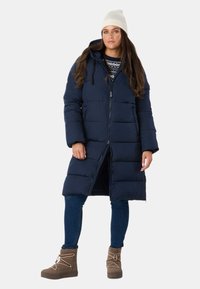 Mørkeblå pufferjakke med lynlås, hætte og frontlommer. Båret over en mønstret sweater, parret med blå jeans og brune ankelstøvler.