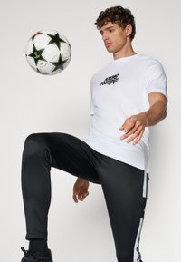 Hvid t-shirt med grafisk print, sorte træningsbukser med sidelommer og sorte sneakers. En fodbold er balanceret på det ene knæ.