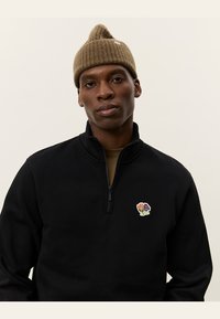Svart zip-up fleece med hög krage, featuring ett färgglatt blommig lappar på vänster bröst. Bärs med en brun stickad mössa.