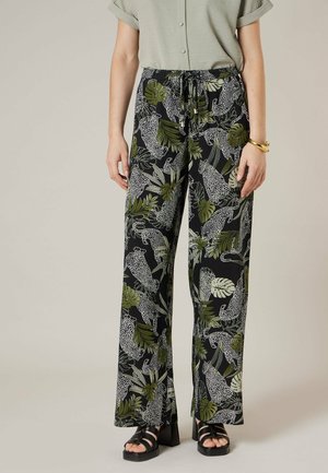 Vrouw draagt zwarte wijde broek met groene tropische bladeren en witte luipaardprint, gecombineerd met zwarte platformsandalen en een groen shirt.