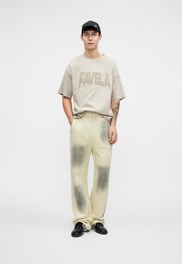 Svjetlosmeđi oversize majica s natpisom "FAVELA", u kombinaciji sa svijetlosmeškim hlačama s tamnijim mrljama. Crna kapa i sjajne cipele upotpunjuju izgled.