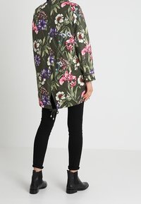 Chemise ample à fleurs vert olive, ornée de fleurs vibrantes roses, blanches et violettes, avec un cordon de serrage à l'arrière. Pantalon noir et bottines.