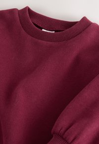 Maglione bordeaux a coste con collo tondo e maniche lunghe, mostrato disteso, che mette in evidenza la trama a maglia e il tessuto morbido.