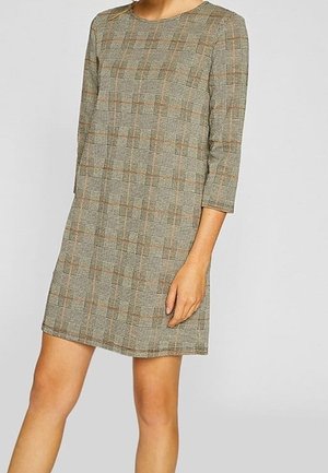 Femme portant une robe à carreaux beige longueur genou, manches longues, avec des accents orange, sur un fond clair uni.