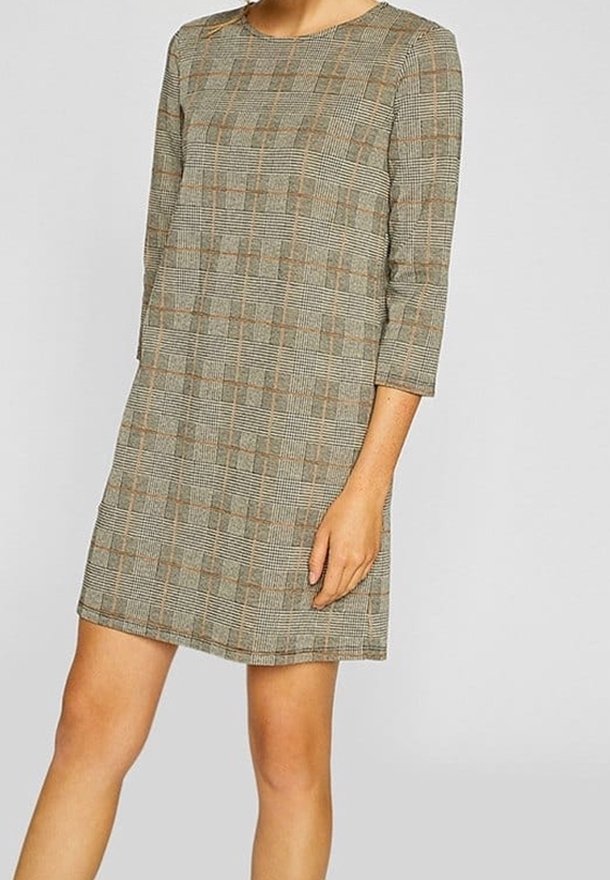Femme portant une robe à carreaux beige longueur genou, manches longues, avec des accents orange, sur un fond clair uni.