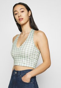 Top crop em gingham verde e branco com decote em V, alças halter e design sem mangas, combinado com jeans de cintura alta azul escuro.