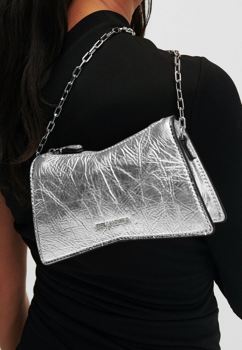 Femme portant un sac à main texturé argent métallique avec une bandoulière en chaîne sur un haut noir à manches longues, montrant la vue arrière et de côté.