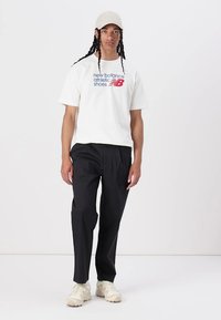 Fehér pamut póló piros és kék "New Balance Athletic Shoes" logóval, fekete nadrággal és krémszínű sportcipővel párosítva.