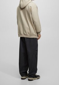 Beżowa bluza z kapturem o luźnym kroju, zestawiona z czarnymi spodniami o szerokich nogawkach. Ciemne sportowe obuwie dopełnia strój. Prosty design, bez widocznych logo.