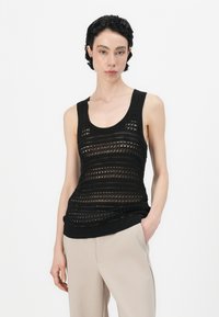 Svart strikket tanktopp med en teksturert, åpen design og ribbede kanter, kombinert med kremfarvede bukser. Enkel bakgrunn.