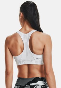 Top sportivo bianco con design a schiena scoperta, caratterizzato da un sottile motivo camouflage e logo sul retro. Texture liscia e forma aderente.