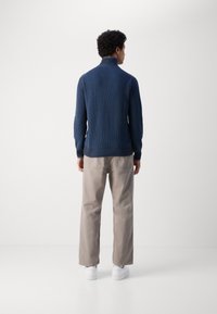 Navy-blauer Strickpullover mit hohem Kragen, kombiniert mit hellbeigen Hosen und weißen Sneakers. Der Stoff ist strukturiert mit gerippten Details.