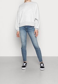 Persona che indossa un ampio sweatshirt grigio chiaro, jeans skinny blu chiaro con zip alla caviglia e sneakers bianco e nere, in piedi su un pavimento grigio.