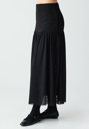 Gonna lunga nera con vita aderente e tessuto trasparente fluente sotto, indossata con collant neri e scarpe Mary Jane nere.