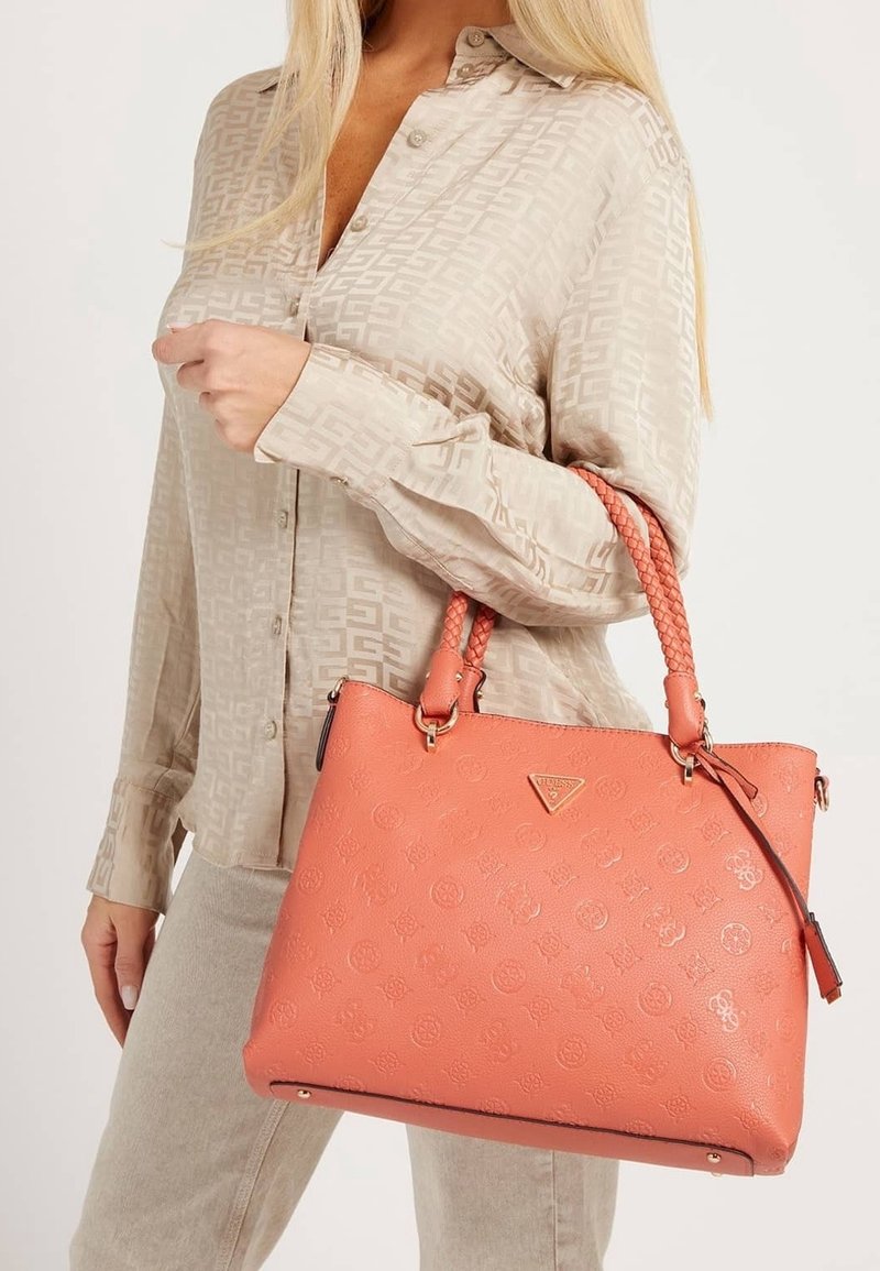 Femme en chemise beige à motifs tenant un sac à main en cuir embossé corail avec des poignées tressées et des accessoires dorés.