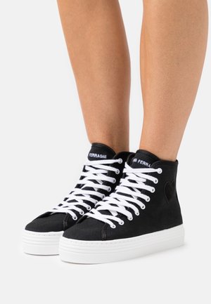 Zapatillas negras de caña alta con cordones blancos y suelas blancas gruesas, usadas sobre piernas desnudas, con un detalle sutil de corazón en el lateral.