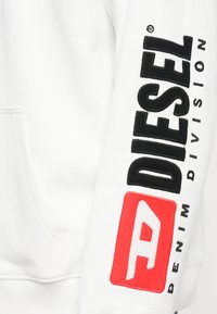 Balts džemperis ar kapuci, uz kura ir melni izšūts "DIESEL" logotips un sarkani akcenti ar tekstu "DENIM DIVISION" uz kreisās piedurknes. Mīksta materiāla tekstūra.