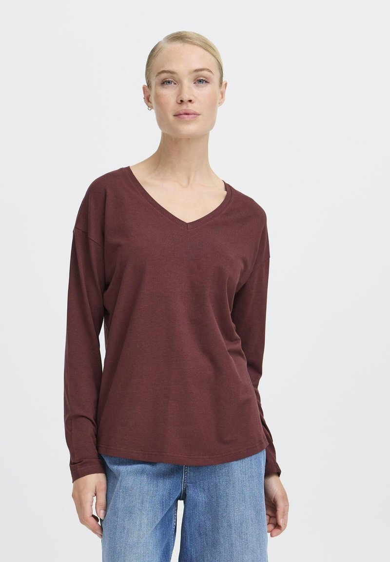 Bordeauxfarbenes Langarmshirt mit V-Ausschnitt aus weichem Stoff, mit lässiger Passform, abfallenden Schultern und abgerundetem Saum. Kombiniert mit blauen Jeans.