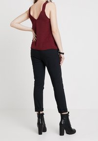 Mujer vestida con una blusa sin mangas de color burdeos, pantalones negros con puños y botines de tacón alto negros con cordones, de pie con una mano en la cadera.