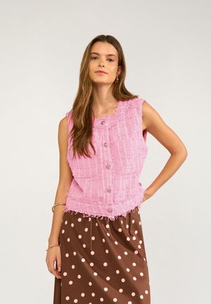 Woman with long brown hair wearing sleeveless pink textured buttoned vest and brown skirt with light pink polka dots, standing against plain background.