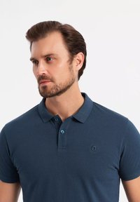 Tummansininen polo-paita, joka on valmistettu tekstuuritusta kankaasta. Paitassa on pieni brodeerattu logo ja kaksipainikkeinen suljenta, jossa on vastakkaiset siniset napit.