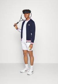 Chaqueta navy con rayas rojas y blancas, camiseta blanca y pantalones cortos blancos. Sosteniendo una raqueta de tenis y una pelota de tenis verde. Zapatillas blancas.