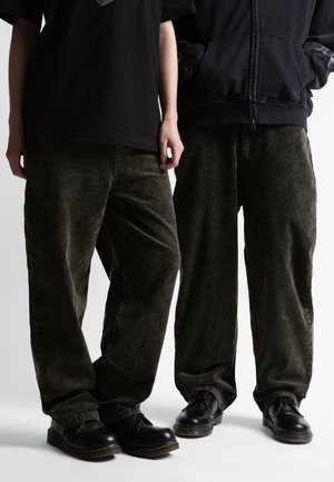 Kaks inimest, kes kannavad tumerohelisi froteepükse ja musti nahast pits-up kingi, üks musta t-särgiga, teine musta lukuga jaskaga.