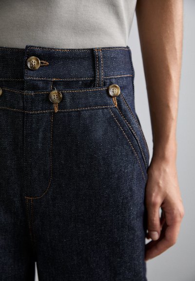 Jean en denim foncé avec une coupe ajustée, présentant deux grands boutons, des coutures orange contrastantes et des poches latérales.