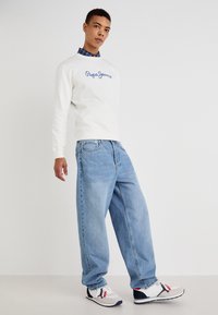 Sudadera blanca con el logotipo azul de "Pepe Jeans", combinada con jeans de pierna ancha de un azul claro y zapatillas deportivas multicolor. Fondo liso.