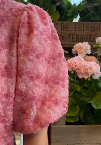 Rosa tyg med texturerade rosor, som har en puffärmad design. Mjukt, genomskinligt material kompletterar gröna lövverk och rosa blommor.