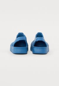Kék slip-on cipők sima műanyagból, nyitott sarokkal, állítható pántokkal és perforált oldalakkal a szellőzés érdekében.