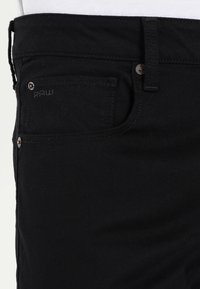 Jeans de denim negro con un corte estándar, bolsillo frontal, logo de la marca sutil y detalles metálicos.
