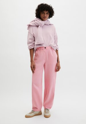 Femme aux cheveux bouclés portant un pull rose clair, un pantalon ample rose, des baskets beiges, et un pull drapé sur les épaules, posant devant un fond blanc.
