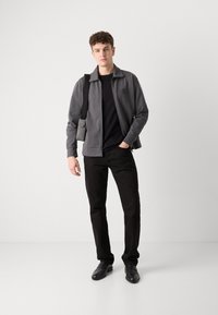 Giacca grigia con zip e una texture liscia, indossata sopra una maglietta nera. Pantaloni neri e scarpe completano l'outfit. Il modello tiene una borsa a spalla.