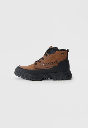 Brown and black ankle boots featuring a suede upper, textured black rubber sole, and lace-up design with fur trim around the collar.