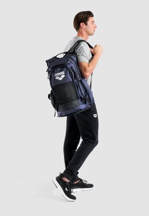 Mochila azul marino con malla negra y logo; cuenta con cremalleras, correas ajustables y un compartimento espacioso. Acentos de tela texturizada.