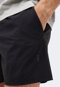 Άνδρας που φοράει μαύρο αθλητικό σορτς με το χέρι στην τσέπη και ένα μικρό λογότυπο κοντά στο τελείωμα στη αριστερή πλευρά.
