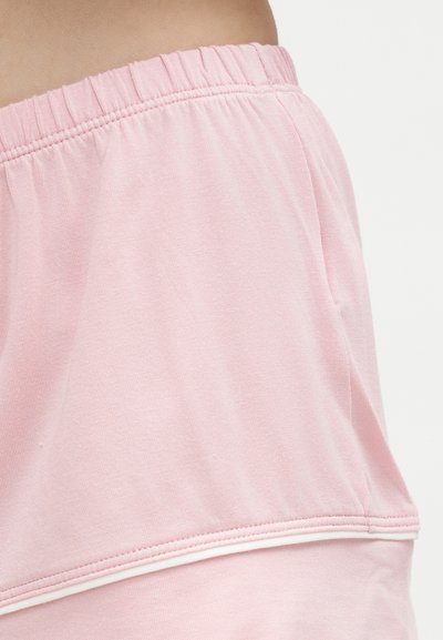 Η μέση φοράει σορτς με ελαστική μέση σε ανοιχτό ροζ χρώμα με πλαϊνή τσέπη και λευκή επένδυση κατά μήκος της ραφής, σε απλό φόντο.