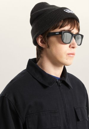 Brunhåret person iført en sort ribstrikket hue og sorte rektangulære solbriller. De har en sort jakke med krave på.