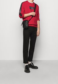 Felpa rossa con logo nero, pantaloni neri, scarpe nere e piccola borsa a tracolla nera. La felpa presenta dettagli a righe testurizzate.