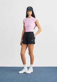 Fiatal nő áll, rózsaszín ujjatlan felsőt, fekete rövidnadrágot, fekete sapkát, fehér zoknit és fehér tornacipőt visel a kék szőnyegen.