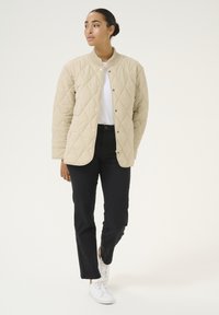 Beige gewatteerde jas met een ronde kraag, drukknoopsluiting en lange mouwen. Gekleed met zwarte broek en witte sneakers op een neutrale achtergrond.