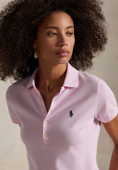 Vrouw met krullend haar die een lichtroze poloshirt draagt met een donkerblauw geborduurd logo en schelpvormige oorbel, kijkt opzij.