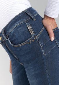 Jeans de mezclilla azul oscuro con costuras naranjas. Presenta una etiqueta metálica que dice "CERISES" en el bolsillo trasero y una textura suave.