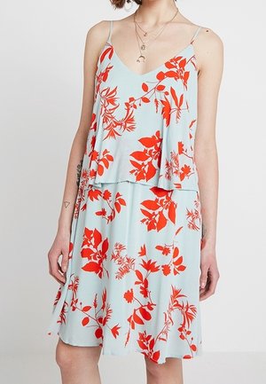 Robe sans manches bleu clair avec imprimé floral rouge vif, col en V, haut à couches, portée par une personne avec des colliers superposés et des tatouages visibles sur les bras.