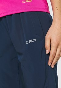 Zblízka záběr na ruku osoby odpočívající na tmavomodrých kalhotách s logem CMP, oblečenou v jasně růžovém topu.