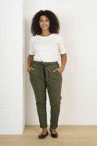 Pantalons ceinturés vert olive avec poches sur les côtés, associés à un haut ajusté blanc à manches courtes. Le mannequin se tient devant un fond neutre.