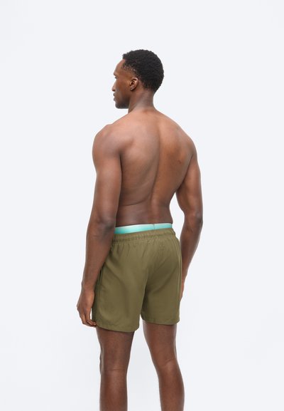 Άνδρας χωρίς πουκάμισο, με την πλάτη γυρισμένη, φοράει χακί σορτς με ανοιχτό μπλε ζώνη μέσης, σε απλό ανοιχτό φόντο.